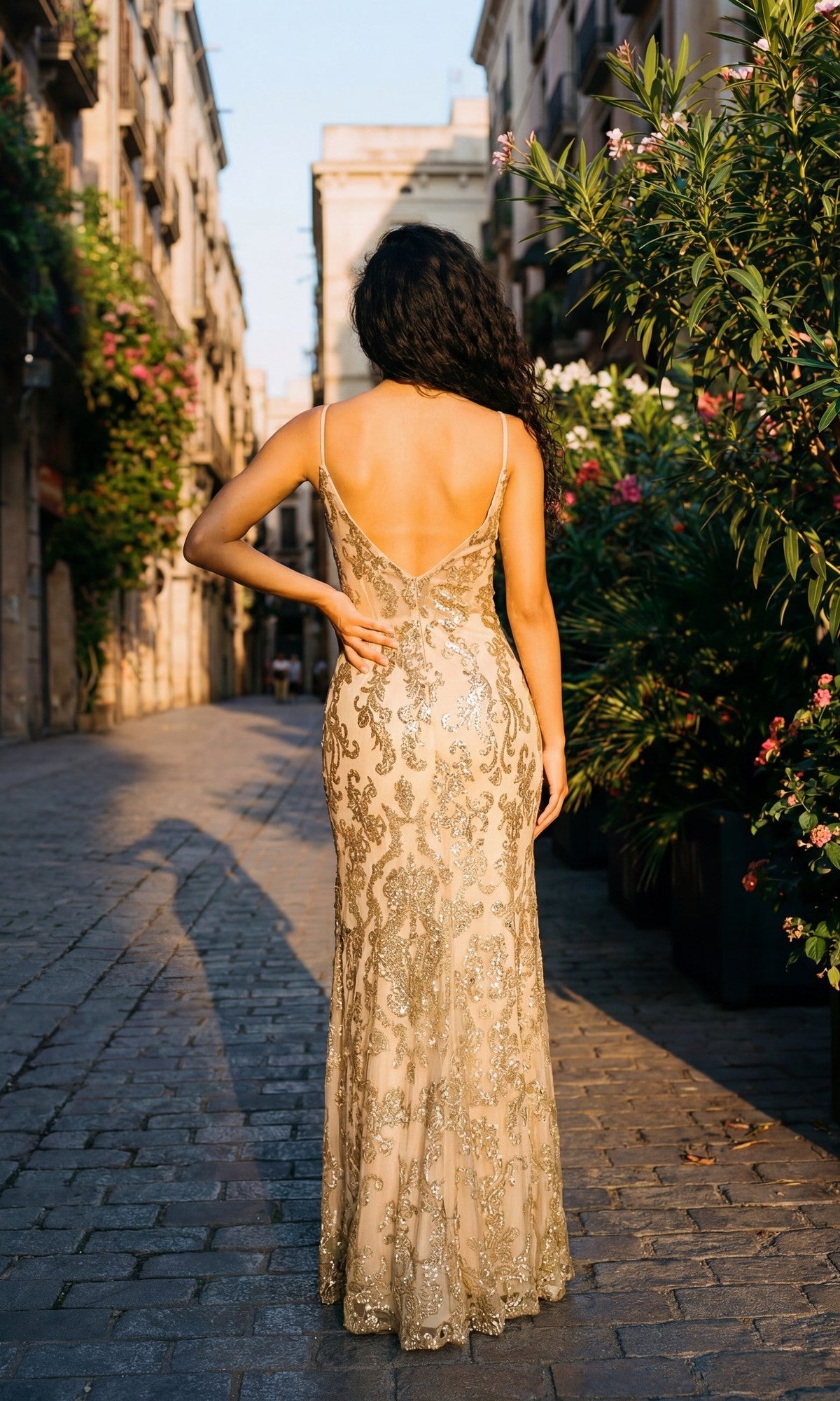 A woman wearing the Chicas 4028 long prom dress, featuring a gold, floor-length sparkly skirt and sheer corset bodice, stands on a cobblestone street lined with plants, facing away from the camera.